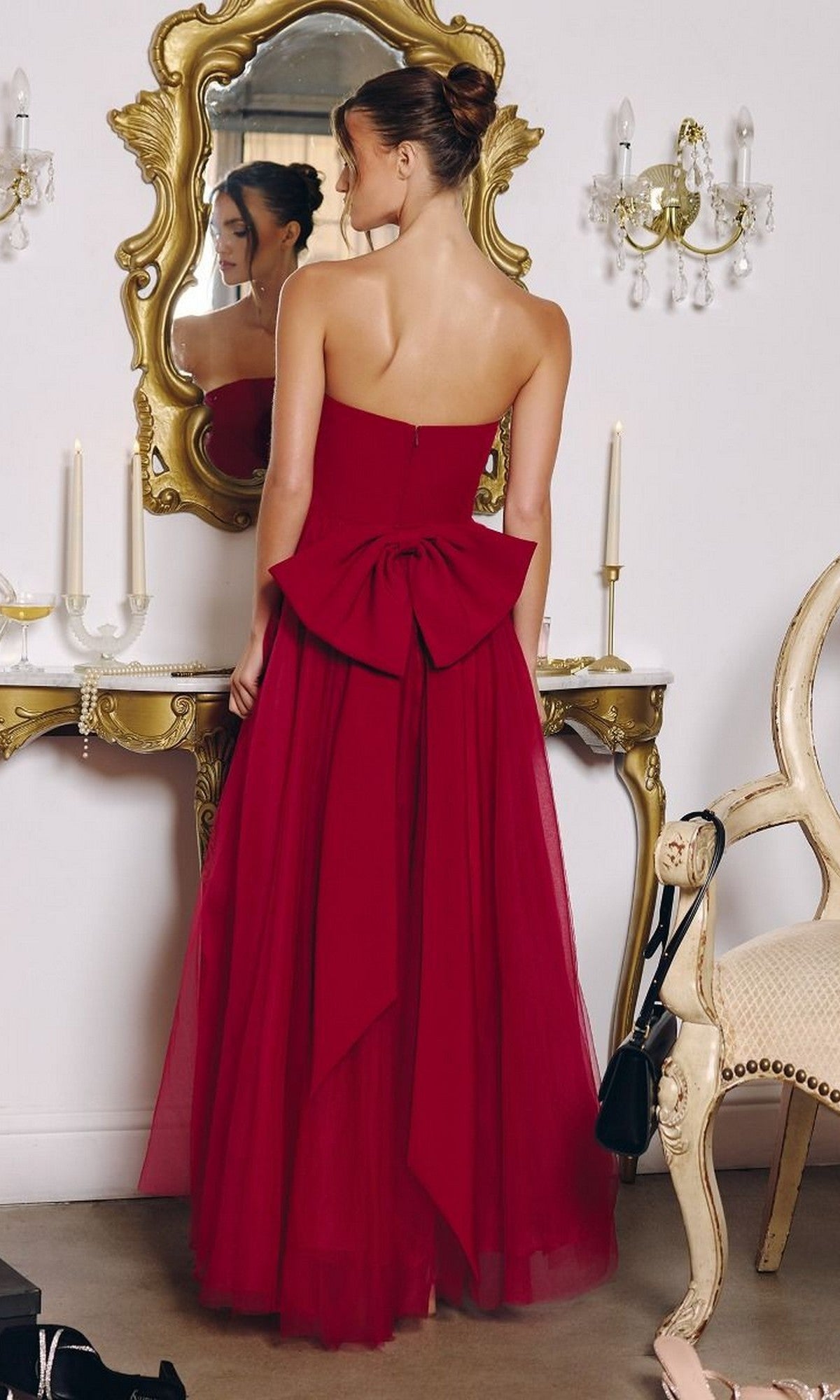 A woman wearing the Classic Strapless Prom Ball Gown: Amoris T712 with a large bow stands before a mirror in an elegant room featuring gold accents, candles, and antique furnishings.